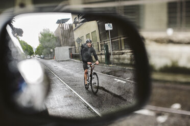 Male commuter riding bicycle on wet street seen through side-view mirror of car - CAVF33072