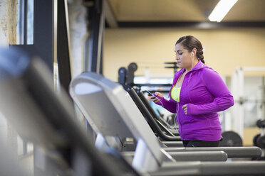 Frau, die ein Mobiltelefon benutzt, während sie im Fitnessstudio auf dem Laufband trainiert - CAVF33085