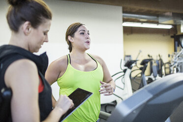 Ausbilder, der einen Tablet-Computer benutzt, während er neben einer Frau steht, die auf dem Laufband im Fitnessstudio trainiert - CAVF33091