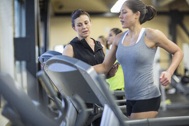 Trainer mit Tablet-Computer instruiert Frau, die auf dem Laufband im Fitnessstudio trainiert - CAVF33094