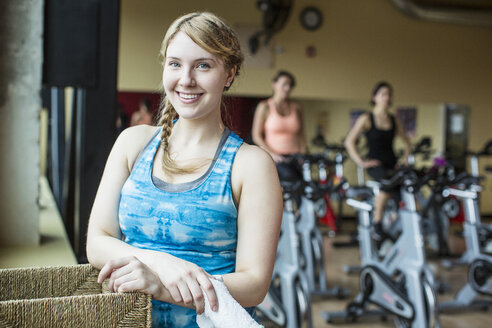 Porträt einer lächelnden Frau mit Freunden, die im Hintergrund im Fitnessstudio trainieren - CAVF33146