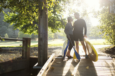 Couple with paddleboards looking at each other while standing on boardwalk - CAVF33336