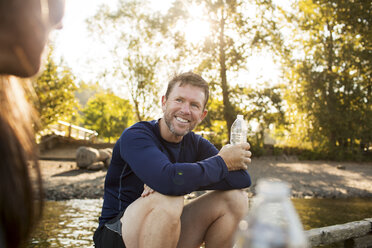 Smiling man holding water bottle while looking at woman sitting on pier - CAVF33349