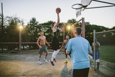 Freunde betrachten einen Mann, der bei Sonnenuntergang auf einem Platz Basketball übt - CAVF33378