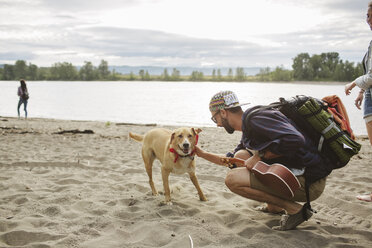 Seitenansicht eines Mannes, der einen Hund streichelt, während er am Flussufer im Sand hockt - CAVF33385