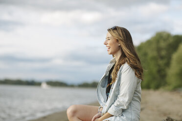 Seitenansicht einer glücklichen Frau, die am Flussufer sitzt - CAVF33388