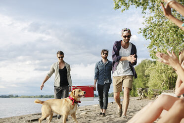 Männliche Freunde tragen eine Kühlbox, während sie auf Sand am Flussufer gegen den Himmel laufen - CAVF33394