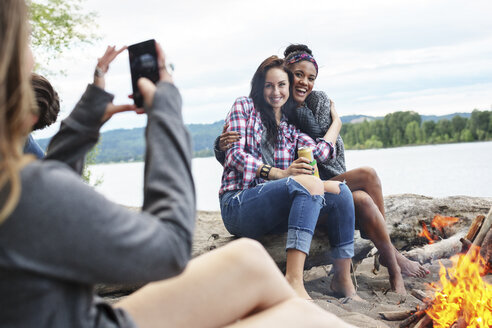 Frau fotografiert glückliche Freundinnen, die auf einem Baumstamm am Lagerfeuer sitzen und gegen den Fluss blicken - CAVF33417