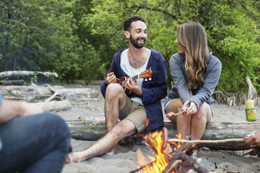 Mann spielt Ukulele, während er mit Freunden am Lagerfeuer am Flussufer sitzt - CAVF33421