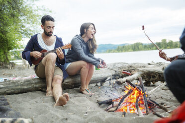 Mann spielt Ukulele, während er mit Freunden zusammensitzt und Würstchen am Lagerfeuer brät - CAVF33423