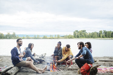 Happy friends roasting sausages on campfire at riverbank against sky - CAVF33425