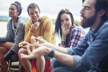 Happy friends holding hotdogs while sitting by river - CAVF33426