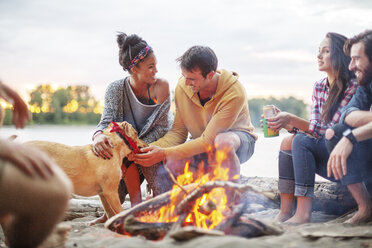 Happy friends with dog by campfire against river - CAVF33432