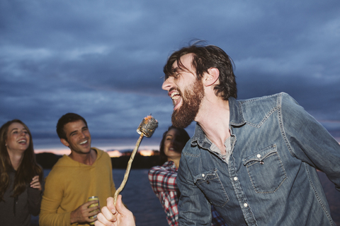 Lächelnde Freunde schauen auf einen singenden Mann, der einen gerösteten Marshmallow auf einem Stock gegen den bewölkten Himmel hält, lizenzfreies Stockfoto