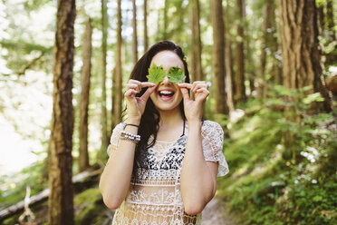 Fröhliche Frau bedeckt Augen mit Blättern im Wald - CAVF33457