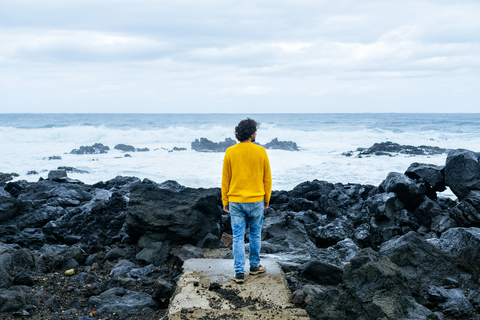 Azoren, Sao Miguel, Rückansicht eines Mannes, der von der steinigen Küste auf das Meer blickt, lizenzfreies Stockfoto