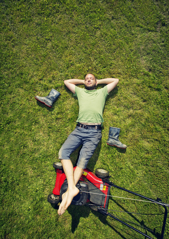 Mann im Gras liegend mit hochgelegten Füßen auf Rasenmäher im Sommer, lizenzfreies Stockfoto