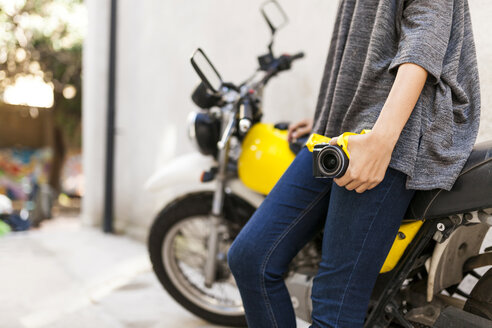 Nahaufnahme einer Frau mit Kamera, die sich an ein Motorrad lehnt - VABF01545