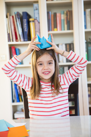 Porträt eines lächelnden Mädchens, das sich zu Hause einen selbstgebastelten Papierhut aufsetzt, lizenzfreies Stockfoto