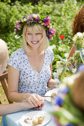 Junge Frau mit Blumenkranz sitzt am Picknicktisch und hält die Hand eines Mannes - FOLF07474