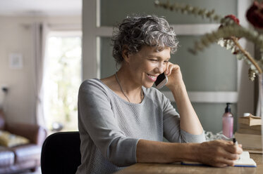 Reife Frau, die zu Hause mit ihrem Smartphone telefoniert und in ein Buch schreibt - CAVF33560