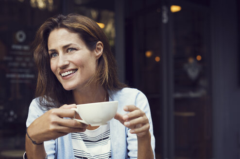 Happy woman looking away while holding coffee cup at sidewalk cafe - CAVF33580