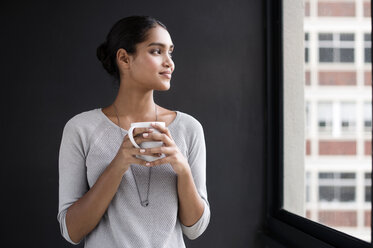 Lächelnde Innenarchitektin, die einen Kaffeebecher hält und durch ein Fenster im Büro schaut - CAVF33617