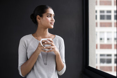 Smiling female interior designer holding coffee mug while looking through window in office - CAVF33617
