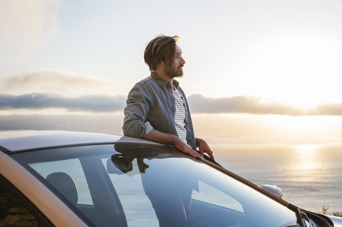 Junger Mann schaut weg, während er bei Sonnenuntergang vor einem Auto am Meer steht, lizenzfreies Stockfoto