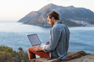 Seitenansicht eines Mannes mit Laptop auf einem Hügel vor einem Berg und dem Meer - CAVF33637