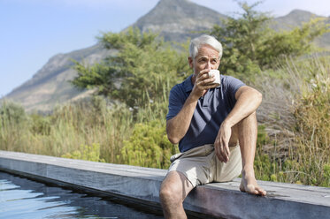 Älterer Mann trinkt Kaffee, während er am Swimmingpool vor einem Berg sitzt - CAVF33643