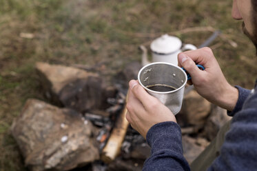 Ausgeschnittenes Bild eines Mannes, der einen Teebecher am Lagerfeuer auf einem Campingplatz hält - CAVF33666