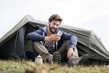 Lächelnder Mann, der ein Smartphone vor einem Zelt auf einem Hügel benutzt, bei klarem Himmel - CAVF33673