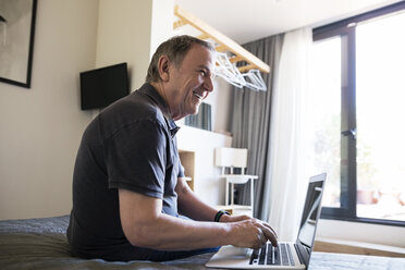 Seitenansicht eines fröhlichen älteren Mannes, der wegschaut, während er im Schlafzimmer einen Laptop benutzt - CAVF33722