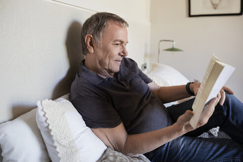 Älterer Mann liest ein Buch, während er zu Hause auf dem Bett sitzt - CAVF33724