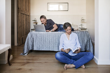 Älteres Ehepaar benutzt Laptop und liest ein Buch im Schlafzimmer - CAVF33729