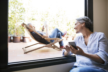 Glückliche Frau schaut auf älteren Mann, der auf einem Liegestuhl auf der Veranda sitzt - CAVF33736