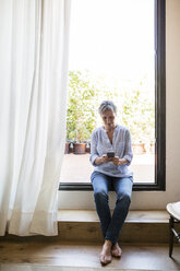 Full length of mature woman using smart phone while sitting on window sill at home - CAVF33739