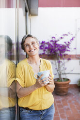 Porträt einer glücklichen reifen Frau, die eine Kaffeetasse hält, während sie auf einer Veranda steht - CAVF33763