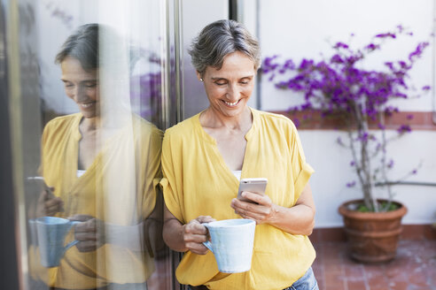 Glückliche reife Frau, die ein Smartphone benutzt, während sie auf einer Veranda einen Kaffeebecher hält - CAVF33764