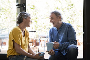 Cheerful senior couple talking while holding coffee mugs by window at home - CAVF33767