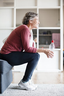 Seitenansicht einer sportlichen reifen Frau, die eine Wasserflasche hält, während sie zu Hause auf dem Sofa sitzt - CAVF33797
