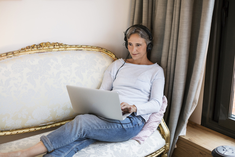 Reife Frau hört Musik und benutzt einen Laptop auf dem Sofa zu Hause, lizenzfreies Stockfoto