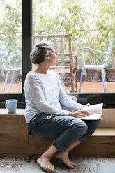 Thoughtful mature woman looking through window while holding book - CAVF33840