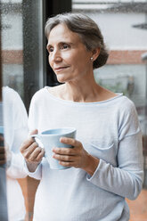 Nachdenkliche reife Frau, die eine Kaffeetasse hält, während sie durch das Fenster zu Hause schaut - CAVF33842