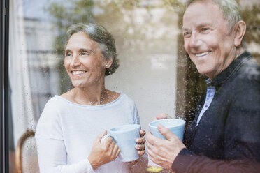 Glückliches älteres Paar, das Kaffeetassen in der Hand hält und durch ein Fenster auf sein Haus schaut - CAVF33844