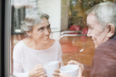 Glückliches älteres Paar, das sich unterhält und dabei Kaffeetassen hält, gesehen durch ein Glasfenster - CAVF33846