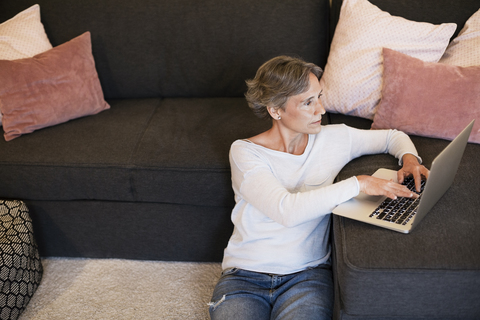 Reife Frau benutzt Laptop, während sie im Wohnzimmer auf dem Boden sitzt, lizenzfreies Stockfoto