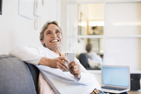 Cheerful mature woman holding smart phone while sitting on sofa at home - CAVF33853