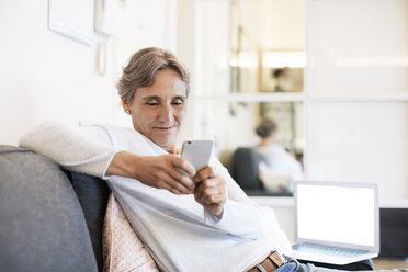 Reife Frau, die ein Smartphone benutzt, während sie zu Hause auf dem Sofa sitzt - CAVF33854